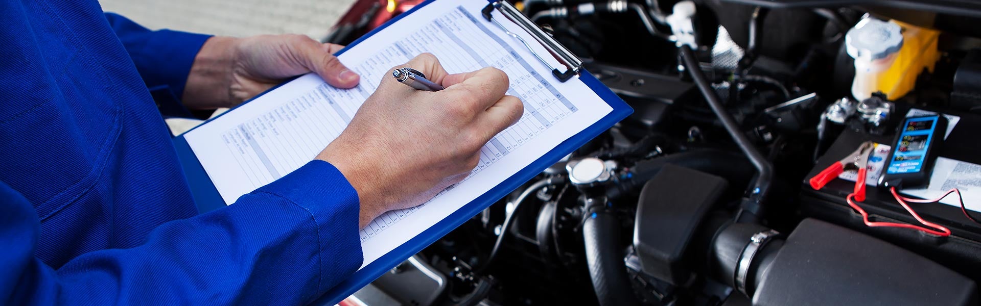 Mechanic filling out a checklist on a clipboard while inspecting an open car engine.